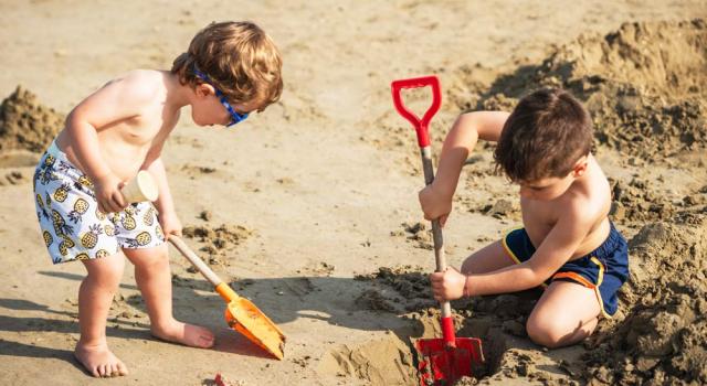 Two joyful kids dig with colorful shovels in the warm beach sand.