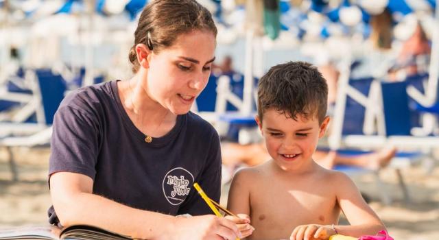 On the beach, a woman helps a child draw, both smiling happily.