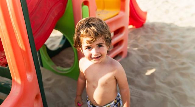 Smiling child plays happily on the sand near a colorful slide.